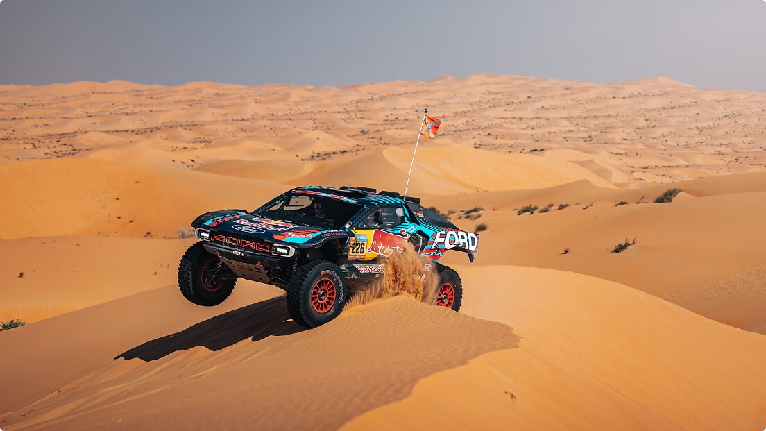 A Ford Performance racing vehicle being driven by a professional race driver over a sand dune in a desert.