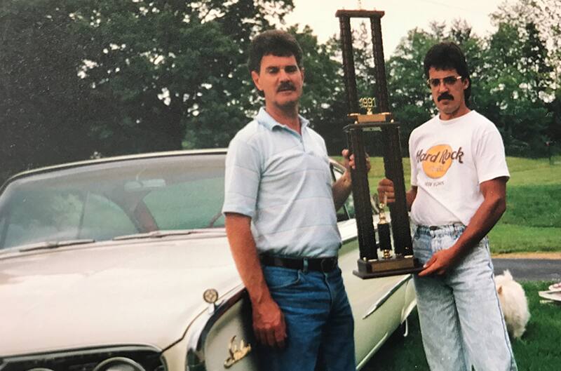 Aged photo of Terry's dad and grandfather holding a trophy next to white Ford Starliner