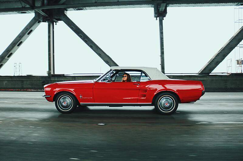 A side view of someone driving a classic red Mustang down the road