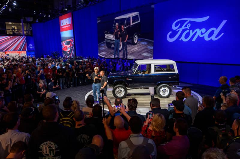 Two men on a stage speaking about a classic blue Bronco