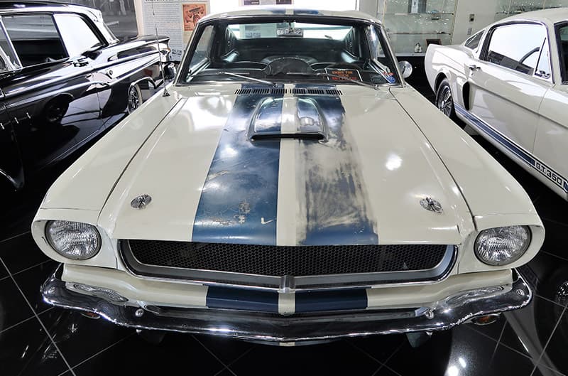 A front end view of a white and blue Mustang on display with a blue stripe rubbing off