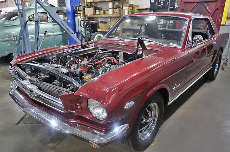 Front of a red Mustang with the hood off and engine exposed in a grarage