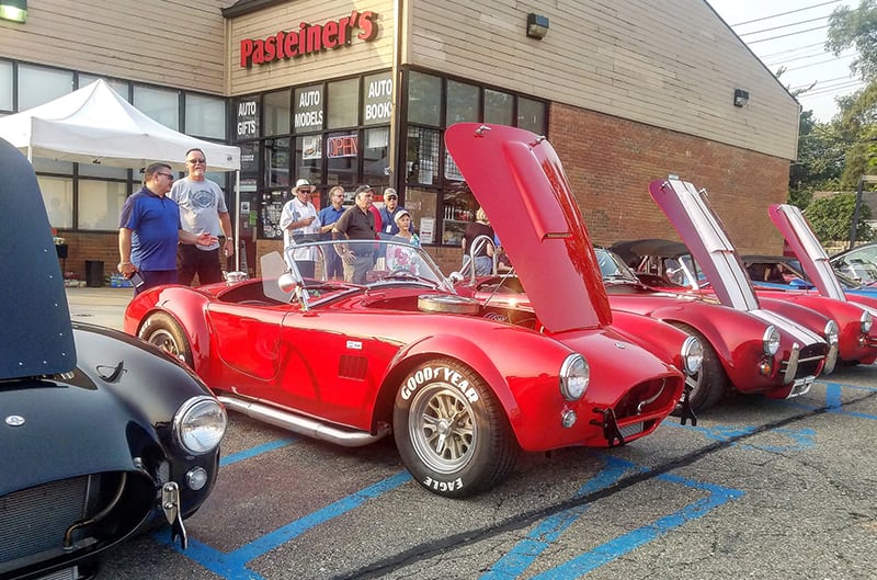 Three red Shelby Cobras parked in a line in the parking lot with hoods up