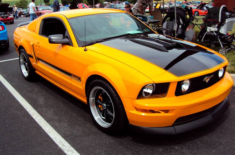 Front profile of a yellow Mach 1 wth a black hood in the parking lot