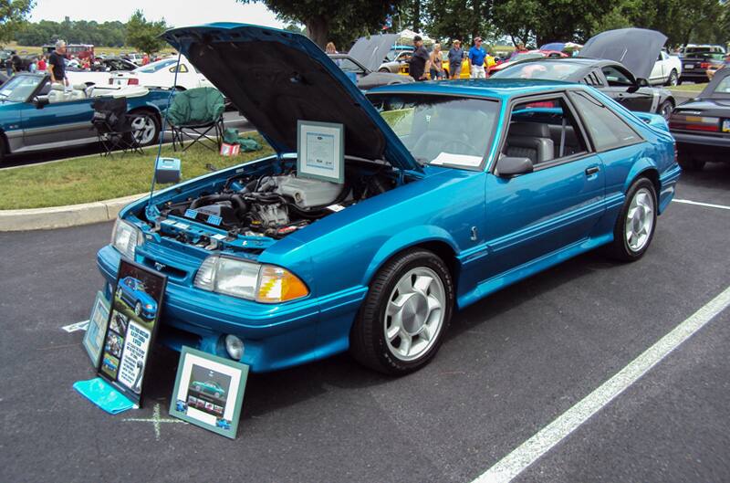 Front profile of a teal blue Mustang with hoodopen in the parking lot