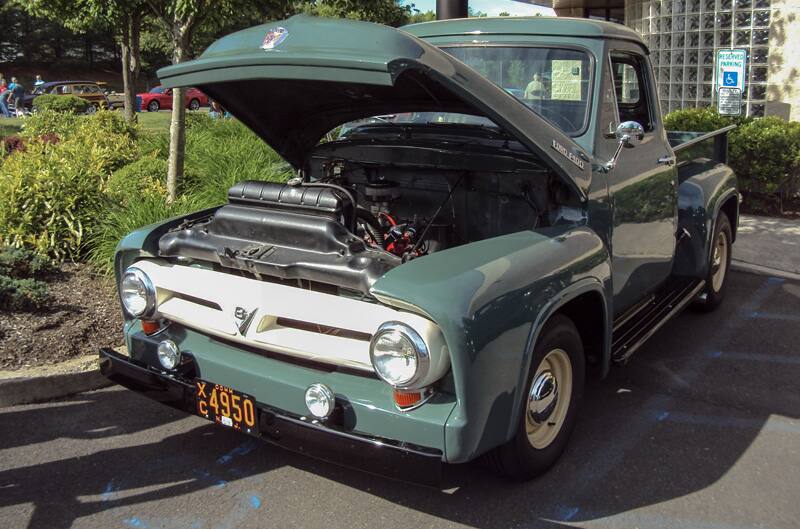 Front of a green classic Ford pickup truck with hood open in parking lot