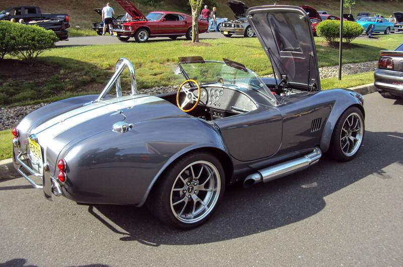 Rear profile of a gray Shelby Cobra with white stripes and hood open in parking lot