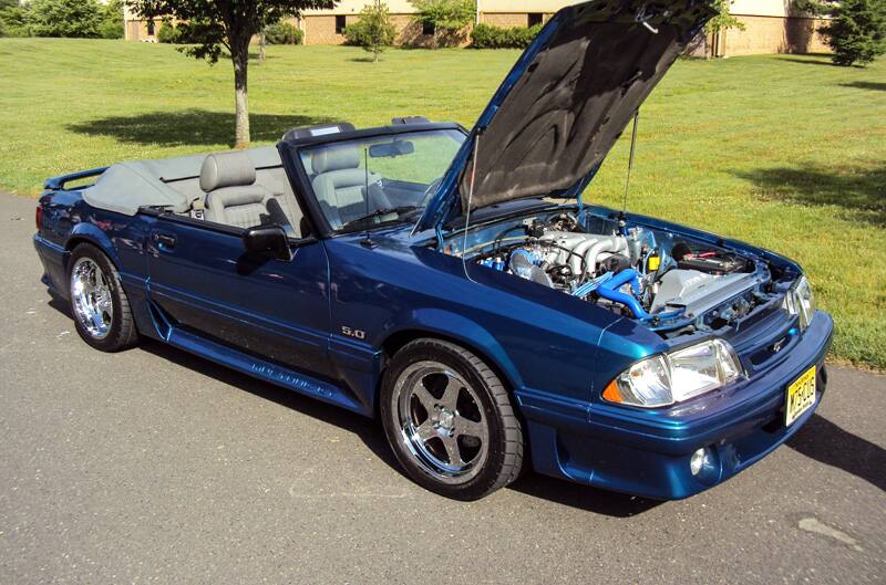 Front profile of a blue Mustang convertible with hood open in parking lot