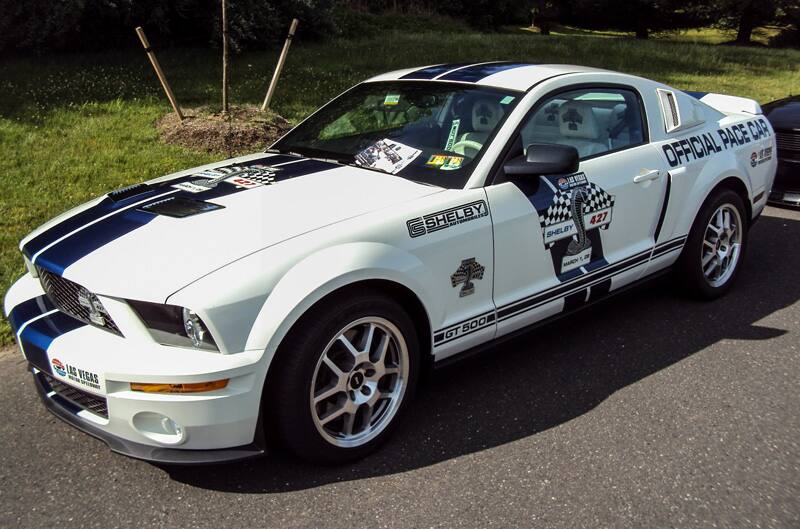 Front profile of a white Shelby GT 500 pace car with blue stripes in parking lot