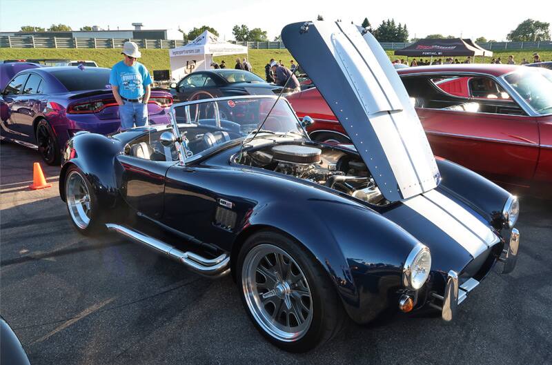 Front profile of a black Shelby Cobra with white stripes and open hood in parking lot