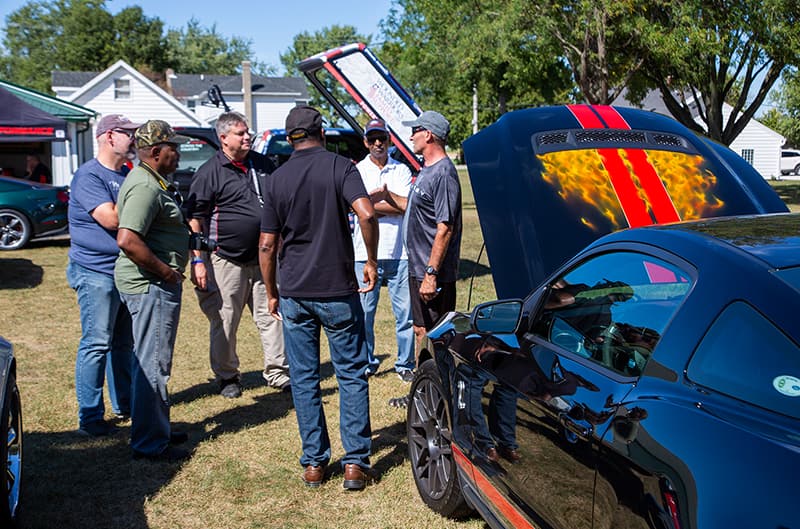 Group of men talking in front of a black Mustang with red stripes on open hood parked on the grass