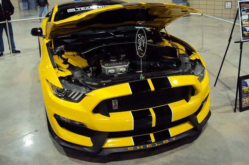 Close up front of a yellow Shelby Mustang with black stripes and open hood on display