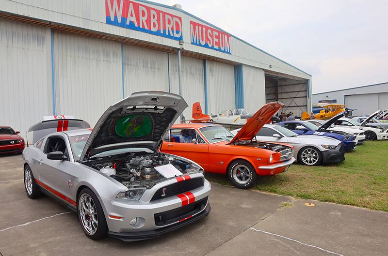 Several mustangs at car show in front of air plane hangar