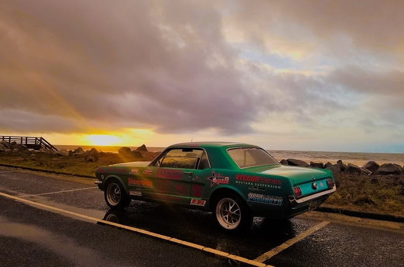Teal Mustang on the road in front of a beach and sunset