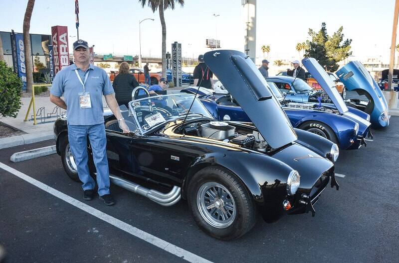 A man is posing next to a black and two blue Shelby Roadsters in a parking lot with the hoods up