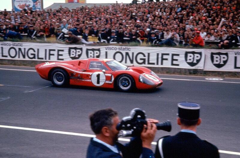 Profile of a orange GT racing on track in front of a crowd of people in the stands