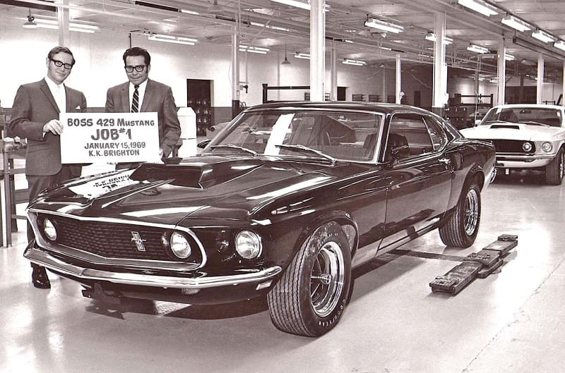 Black and white image of two men holding up Boss 249 Mustang poster in front of a front profile of a Musutang in a garage