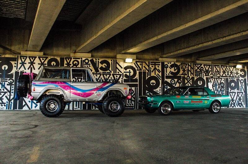 Gray pink and blue Bronco facing a teal Mustang under a bridge