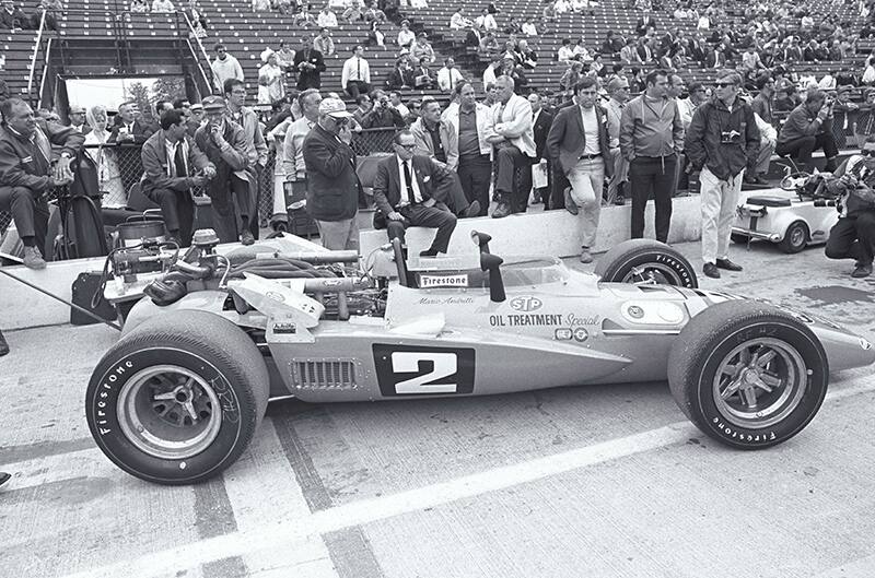 Black and white photo of profile of indycar at the track with people surrounding