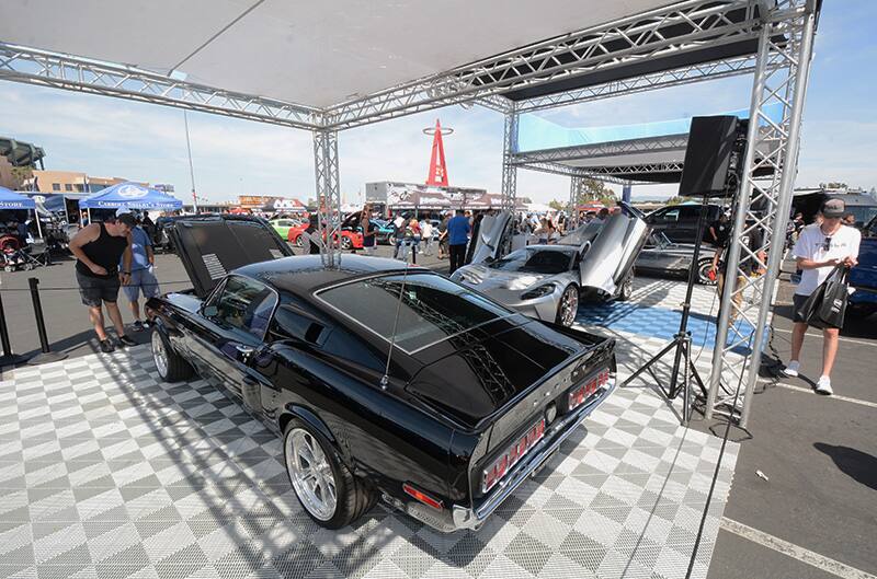 Rear of a black Mustang on display under a tent with people looking under the hood