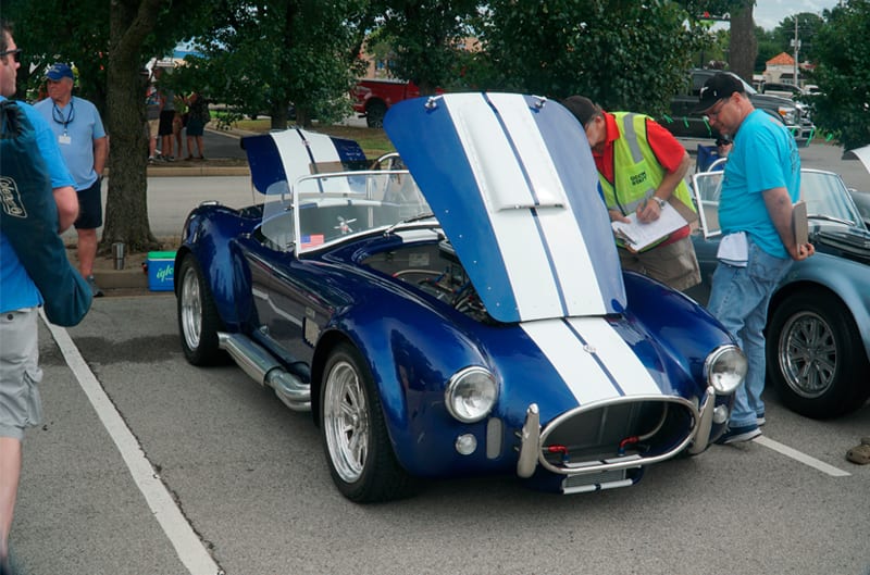 Front of blue Shelby Roadster with white stripes with hood and trunk open in parking lot with people observing