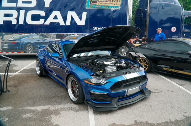 Front profile of blue Shelby Mustang with hood open in parking lot in front of Shelby American semi truck