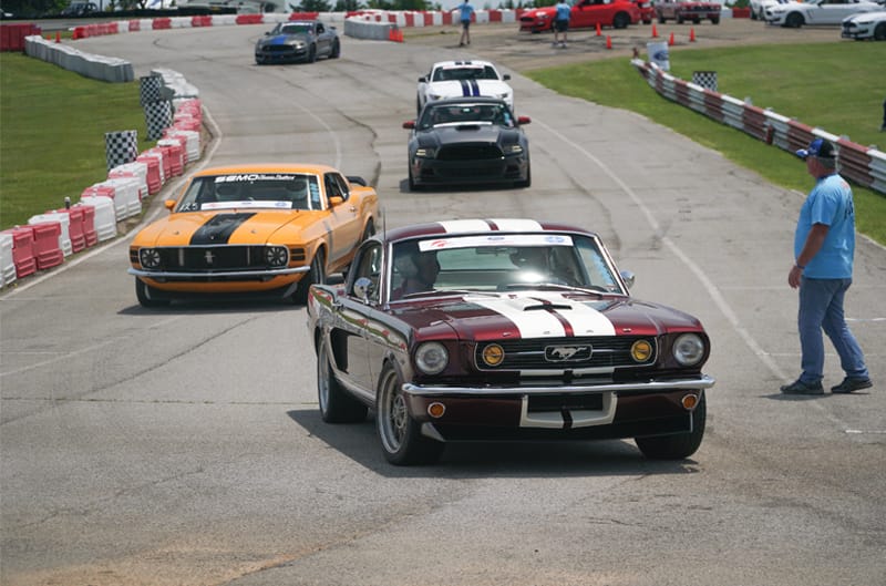 Front facing red Mustang with white stripes leading a pack of Mustangs on the track