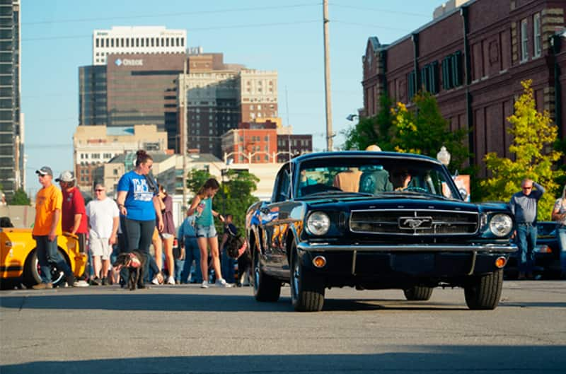 Front of a black Mustang in the parking lot in front of people and a city view