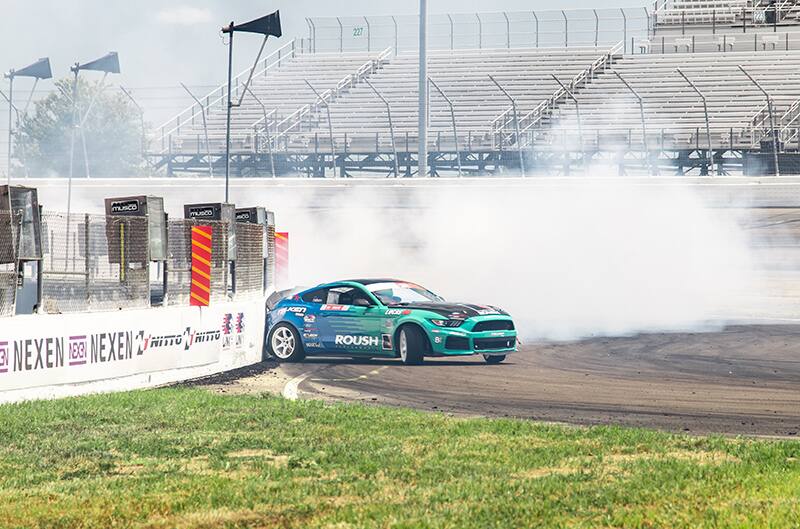 Profile of Justin Pawlak drifting his blue and green Mustang on the track near a wall