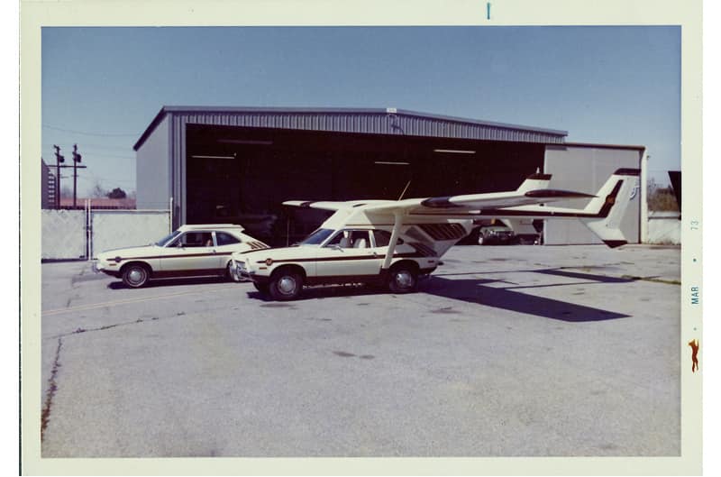 Vintage photo of the white Galpinized Flying Ford Pinto in front of a garage and another white Ford Pinto behind it