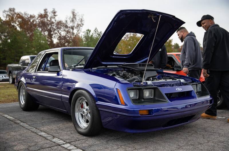 Front profile of blue Mustang with hood open and two men looking at it in parking lot