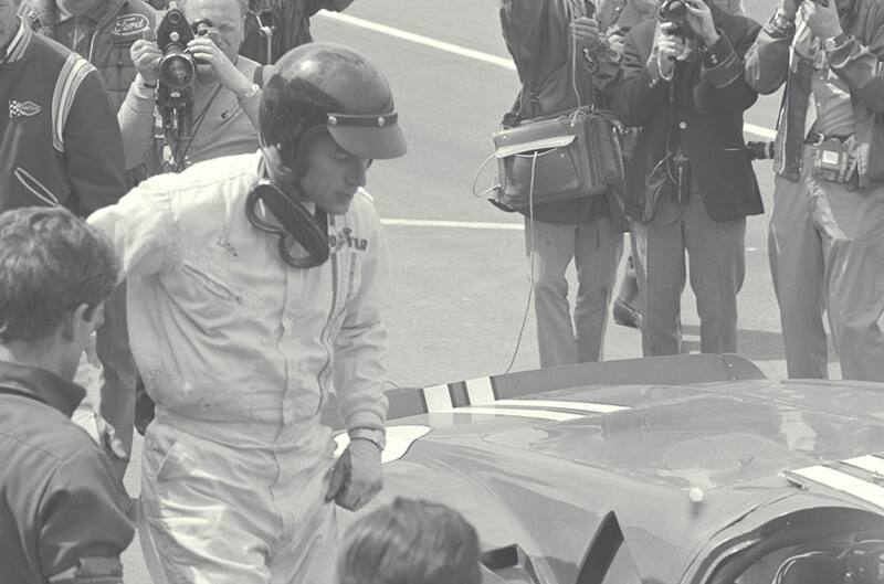 Black and white photo of Dan Gurney walking up to GT on the track with photographers behind him