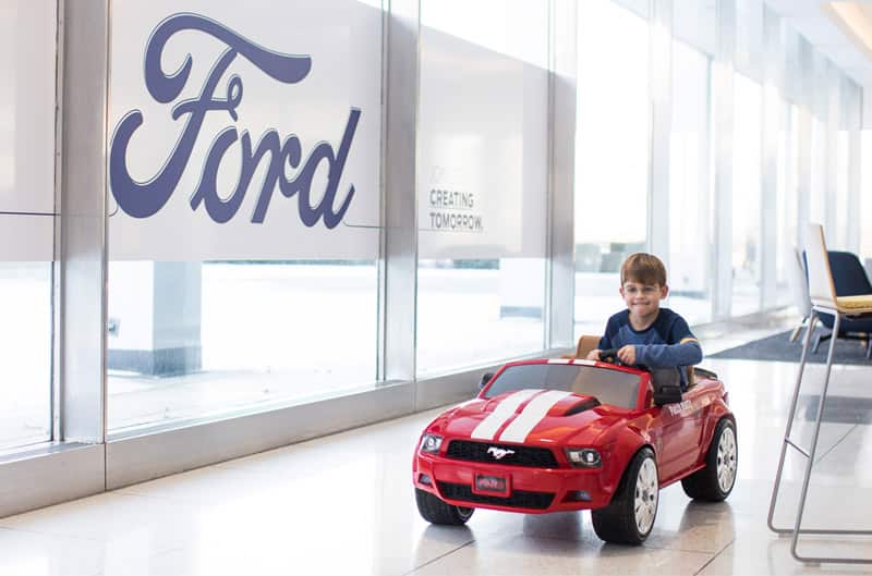 Patch Hurty driving a toy red Mustang inside a Ford dealership