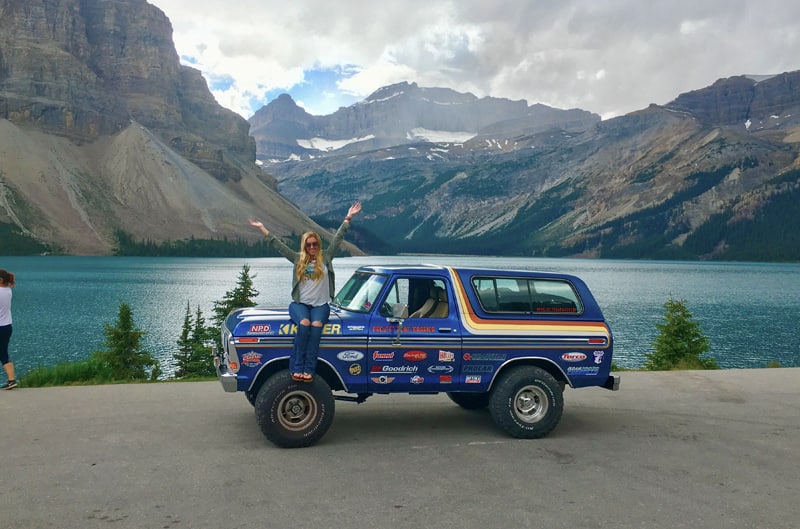 Profile of blue Bronco with Courtney sitting on the hood in front of Artic Ocean and mountains