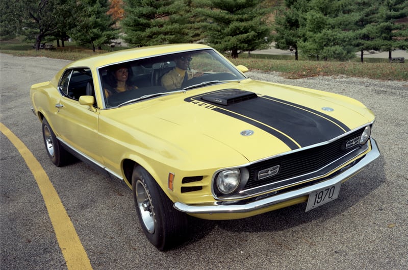 Front of yellow Mustang Mach 1 with black stripe on hood driving on the road