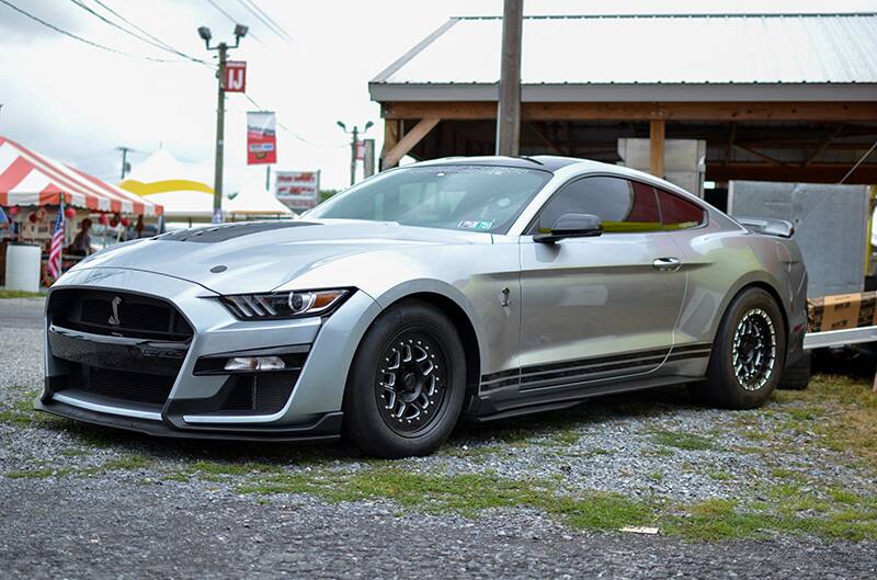 Front profile of a silver Shelby Mustang parked on the grass in front of tents and a pavilion