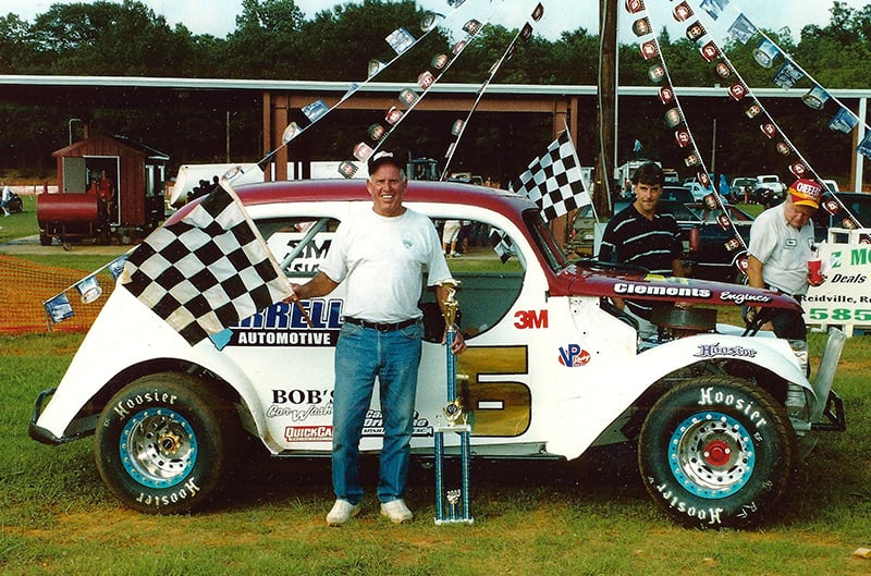 Larry posing in front of white and red Ford race car holding trophy and flag