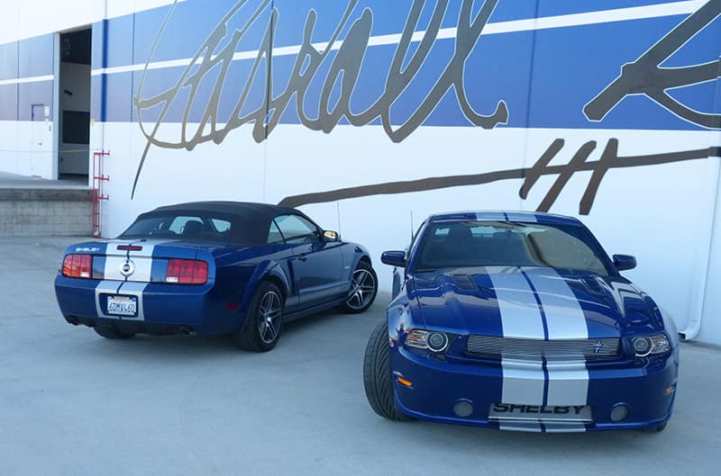 Blue Mustangs with silver stripes parked in from of building with Carroll Shelby signature painted on side