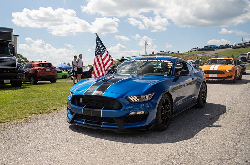 Blue GT350 Mustang with american flag hanging from passenger window driving at carlisle