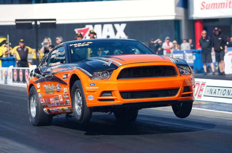 With front wheels up in the air, a black and orange Cobra Jet Mustang races down dragstrip where fans can be spotted in the background