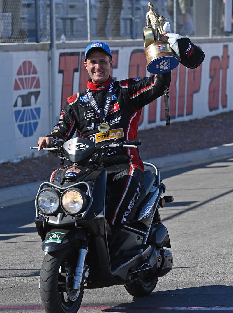 Bob Tasca III ridingo n a motorcycle down the drag strip holding trophy