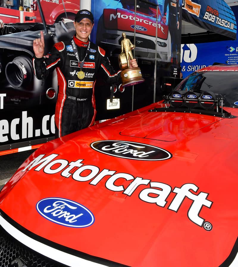 Close up of Bob Tasca III standing behind front of NHRA Nitro Funny Car Mustang holding up trophy