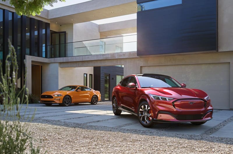 Front of a red Mustang Mach E and a yellow Mustang in the driveway of a modern home