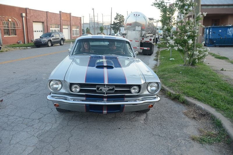 Two people inside a silver and blue classic Ford Mustang driving down the road 
