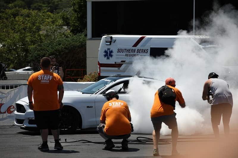 A white Ford Mustang burning out while staff members are recording 