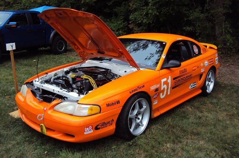 A front side view of an orange Ford Mustang on display with the hood up 