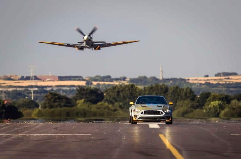 A picture of the Ford Eagle Squadron Mustang GT driving on the road while an Eagle Squadron Spitfire is flying in the back