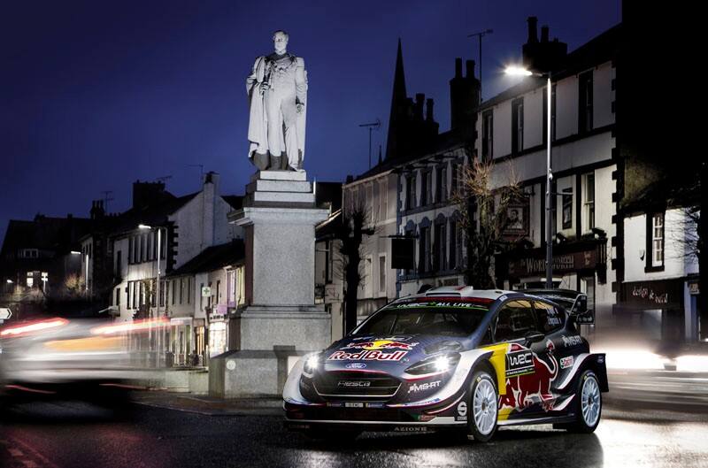 A Red Bull Ford Fiesta zooming around a statue on a road